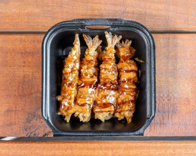 Four crispy tempura shrimp drizzled with creamy and dark sauces in a black takeout container.