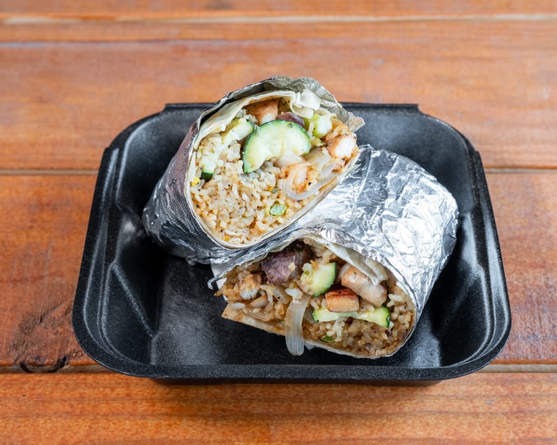Two foil-wrapped burritos, cut in half, showing rice, zucchini, and meat, in a black takeout container.