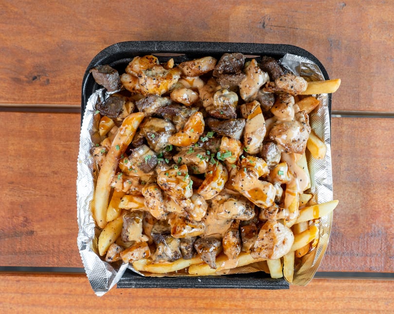 Loaded fries with chicken, steak, creamy sauce, and chives in a foil-lined container.