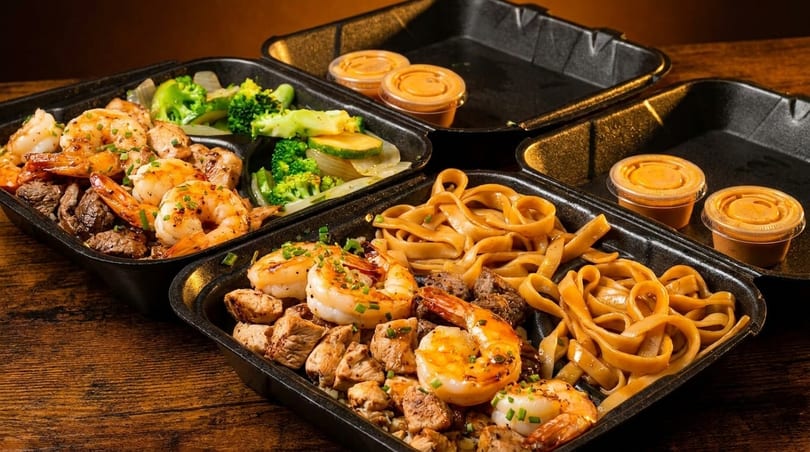 Two takeout containers with shrimp, chicken, steak, noodles, and vegetables, plus sauce cups.