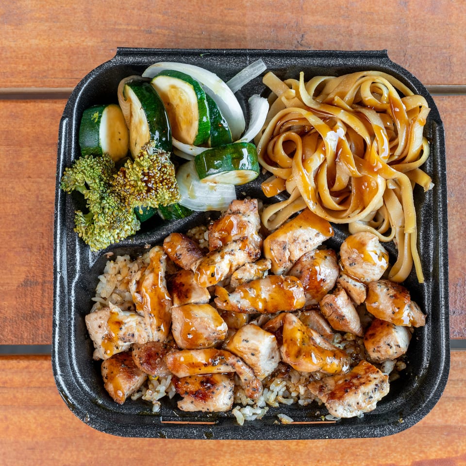 Black takeout container filled with chicken, rice, noodles, and steamed vegetables on wood.