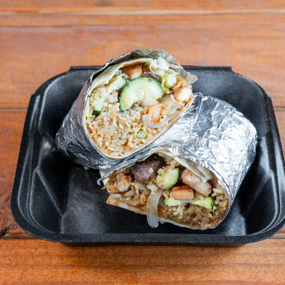 Two foil-wrapped burritos, cut in half, filled with rice, zucchini, and meat in a black takeout container.