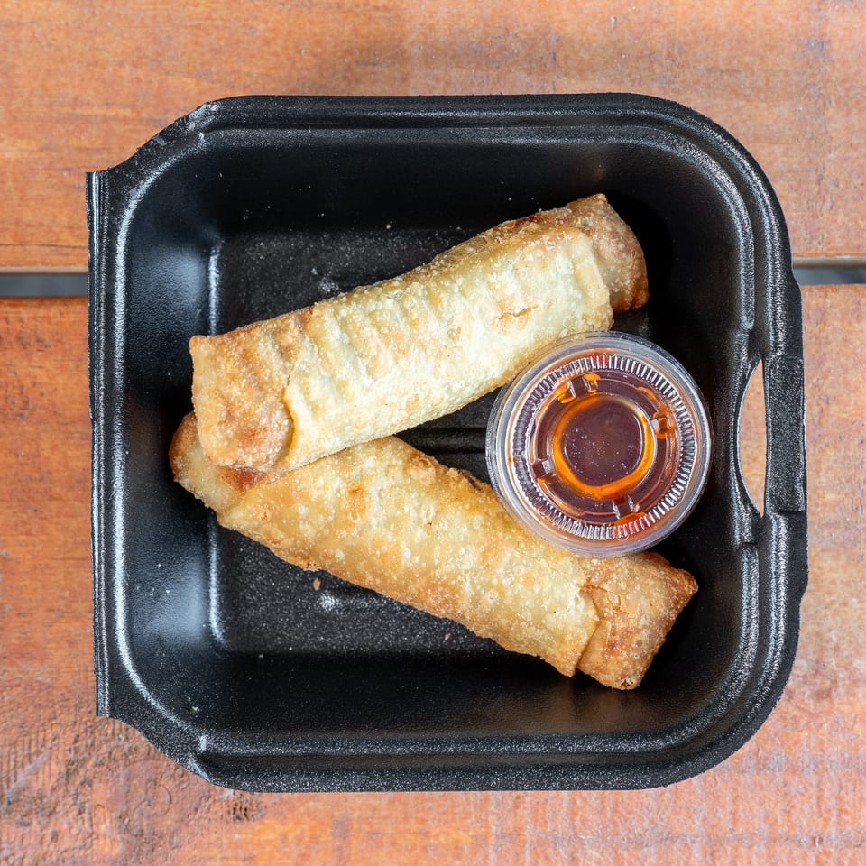 Two crispy egg rolls with dipping sauce in a black to-go container on wood.