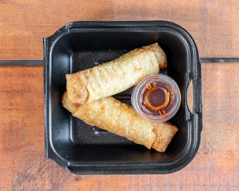 Two crispy egg rolls with dipping sauce in a black to-go container on wood.