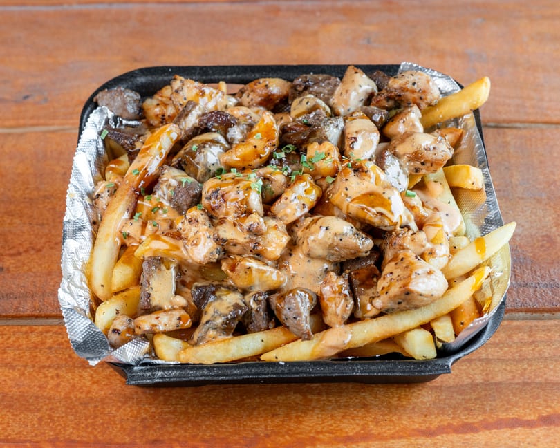 Golden french fries, chicken, and beef pieces covered in creamy sauce and chives in a black container.