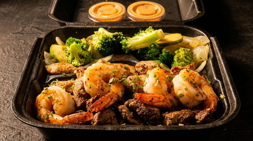 Shrimp, steak, and chicken with broccoli, zucchini, and onions in a black takeout container.